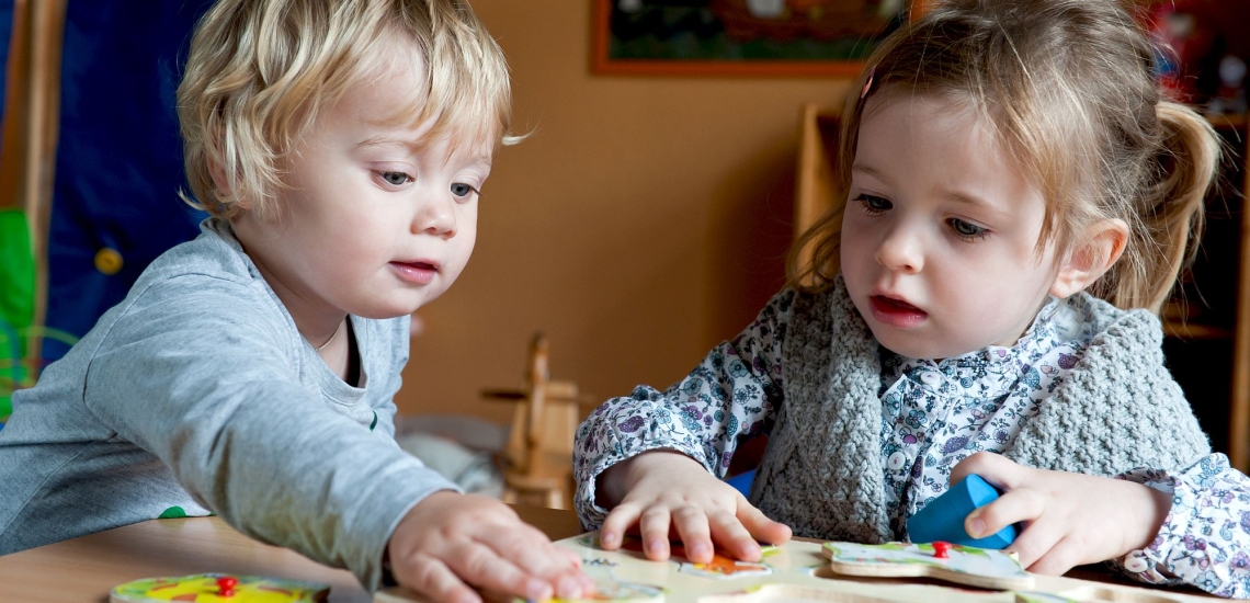 Zwei Kleinkinder sitzen an Tisch und spielen mit Holzpuzzle