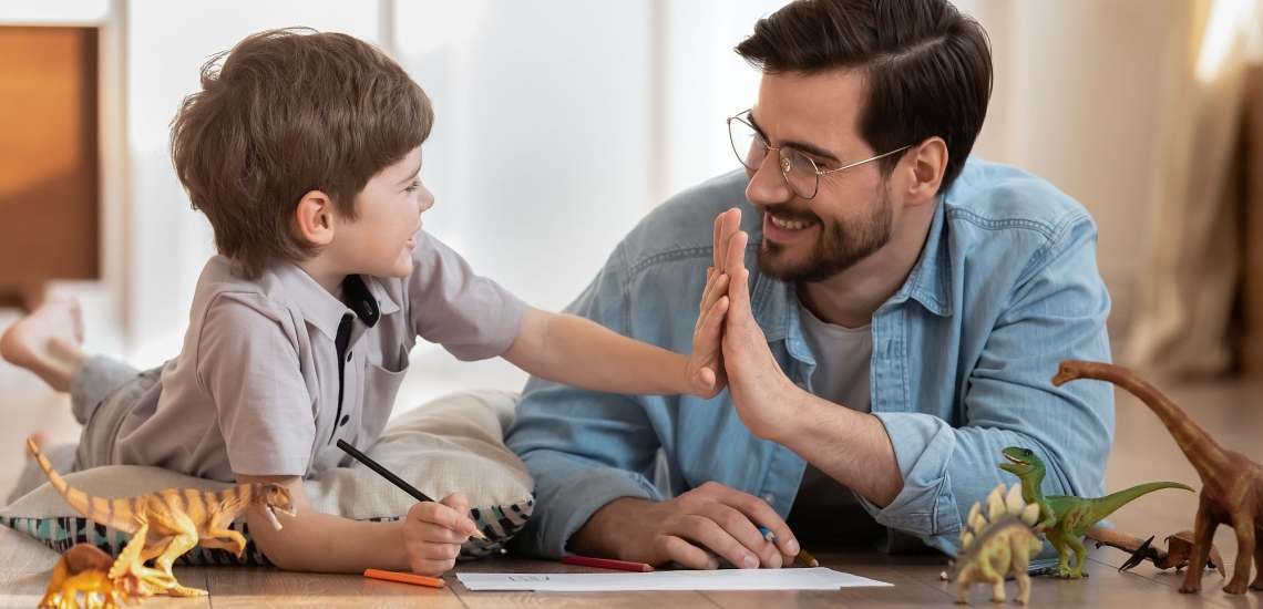 Vater und Sohn liegen lachend mit Papier, Stiften und Dino-Figuren auf dem Boden und machen High five