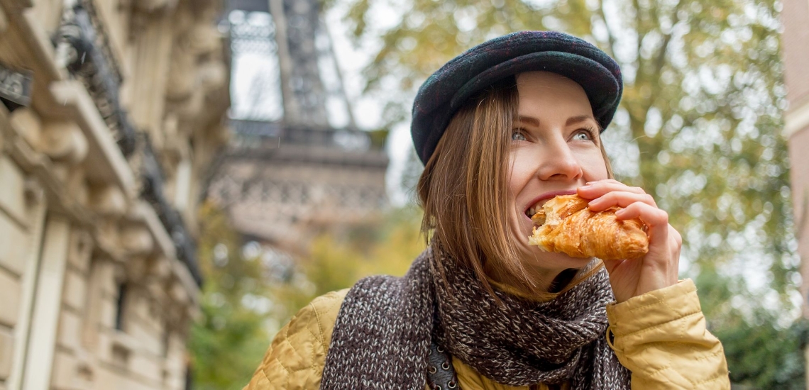 Frau mit Baskenmütze und halblangen dunkelblonden Haaren beißt in Croissant vor Eiffelturm