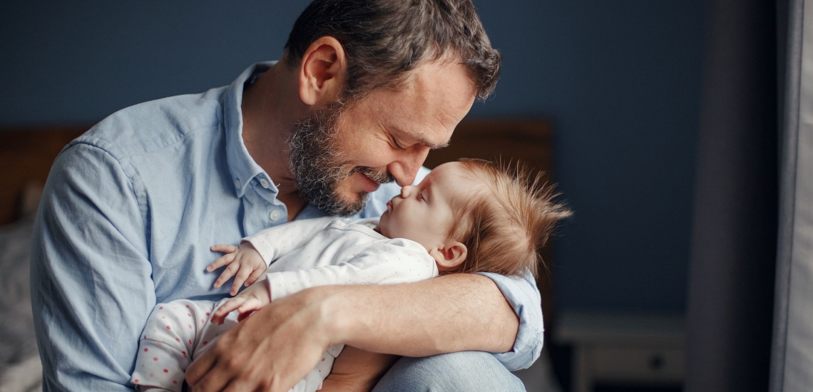 Vater hält Baby liebevoll im Arm und stupst seine Nase an die Nase des Babys.