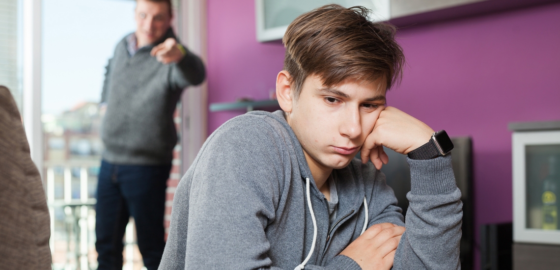 Vater steht im Hintergrund und Zeit aufgebracht mit dem Finger auf den jugendlichen Jungen, der ihm den Rücken zudreht.