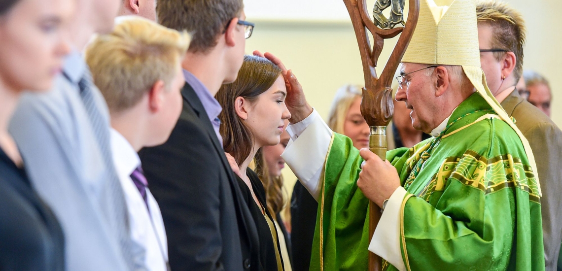 Ein Bischof zeichnet mit Chrisamöl ein Kreuz auf die Stirn eines Firmlings, der in einer Reihe mit anderen Firmlingen steht