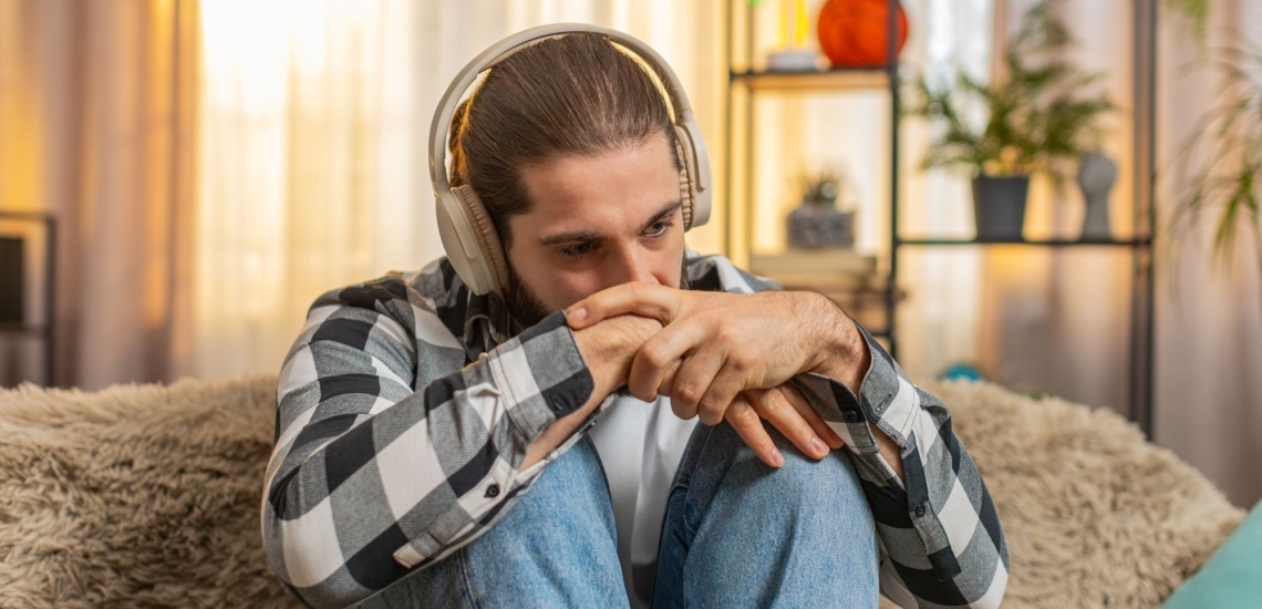 Jugendlicher mit zusammengebundenen Haaren, Karohemd und Kopfhörern sitzt traurig und in sich gekehrt auf dem Sofa