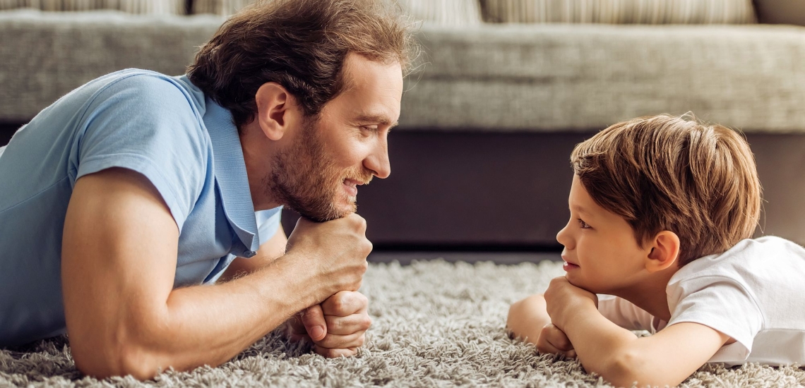 Vater und Kind liegen sich auf dem Wohnzimmerteppich gegenüber und schauen sich an