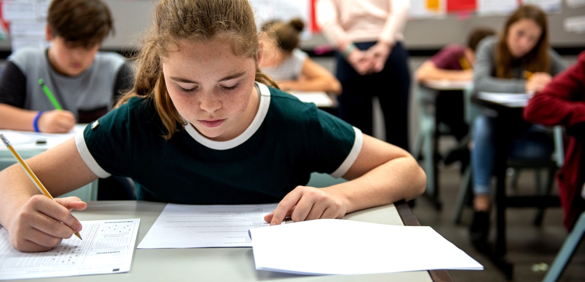 Eine Jugendliche mit grünem Shirt sitzt an einem Tisch im Klassenraum einer Schule und schreibt einen Test. Im Hintergrund sind Mitschülerinnen und Mitschüler in der gleichen Situation zu sehen.