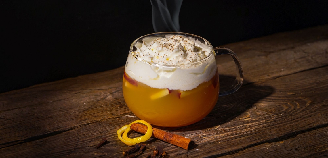 Dampfender Punsch in gläserner Tasse mit Sahne auf Holztisch, daneben Zimtstange und Zitronenschalenring