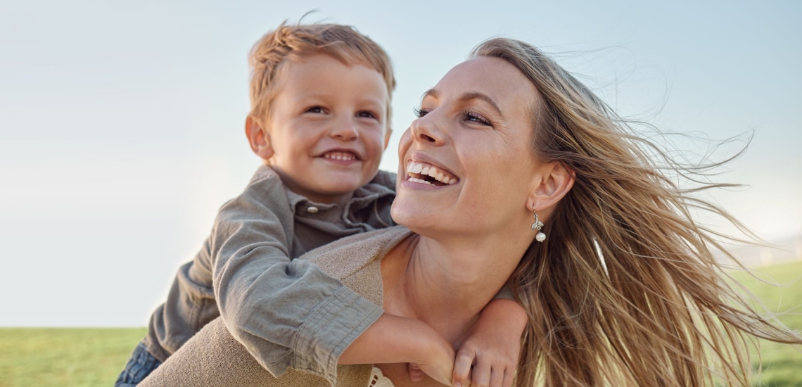 Mutter trägt Kind auf dem Rücken fröhlich im Sommer mit Wind im Haar