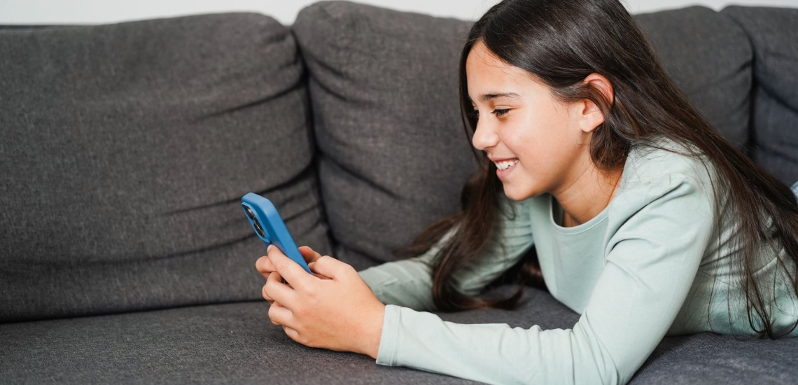 Kind mit langen Haaren liegt fröhlich auf Sofa und schaut in Handy