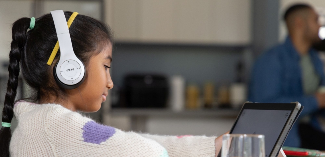 Kind sitzt in der Küche mit Kopfhörern am Tablet, Vater steht im Hintergrund