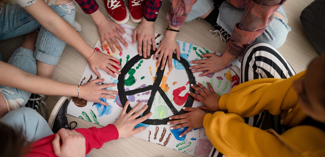 Hände von Jugendlichen liegen auf einem Plakat, auf dem ein Peace-Zeichen gemalt ist und bunte Handabdrücke zu sehen sind.