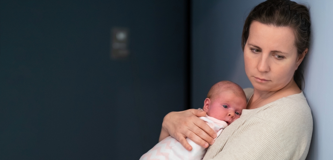 Eine Frau blickt nachdenklich auf den Boden. Dabei hält sie ein Baby auf dem Arm.