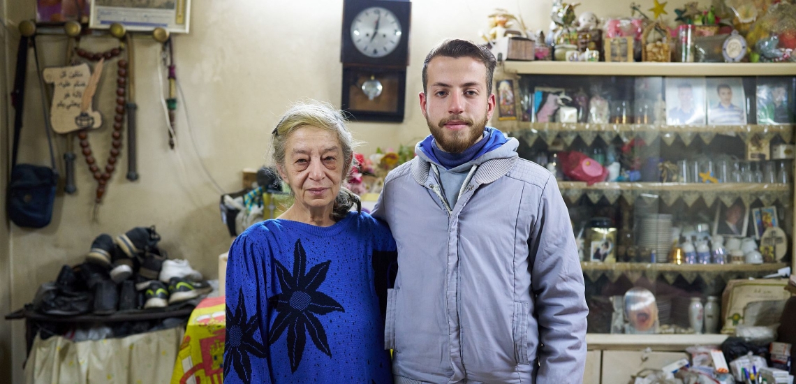 Ältere Frau mit jungem erwachsenen Sohn in Wohnung vor Wand mit Regal und Uhr