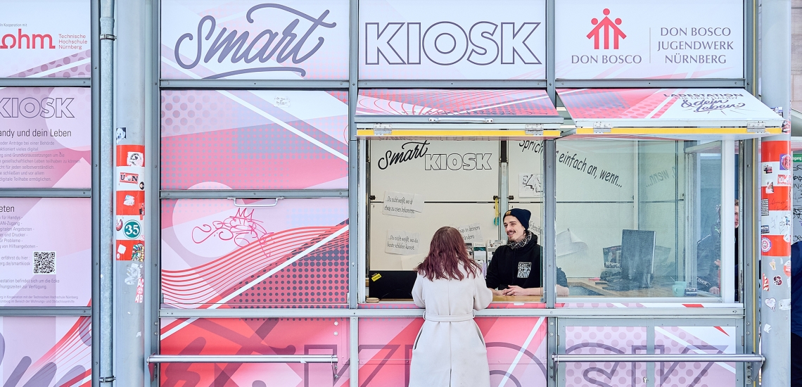 Eine junge Frau steht am Smart Kiosk und unterhält sich mit einem jungen Mann, der Kaffee ausgibt.