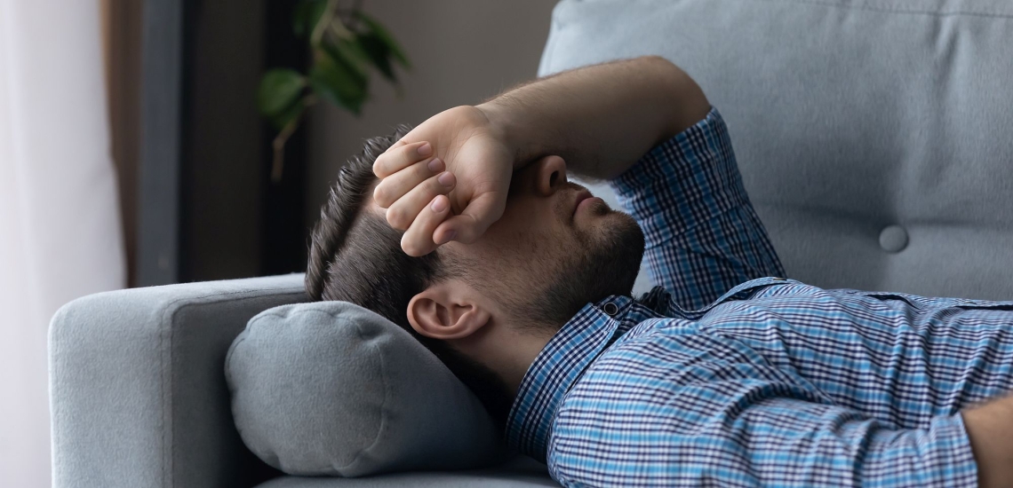 Mann liegt krank auf Sofa und hat den Arm über das Gesicht gelegt