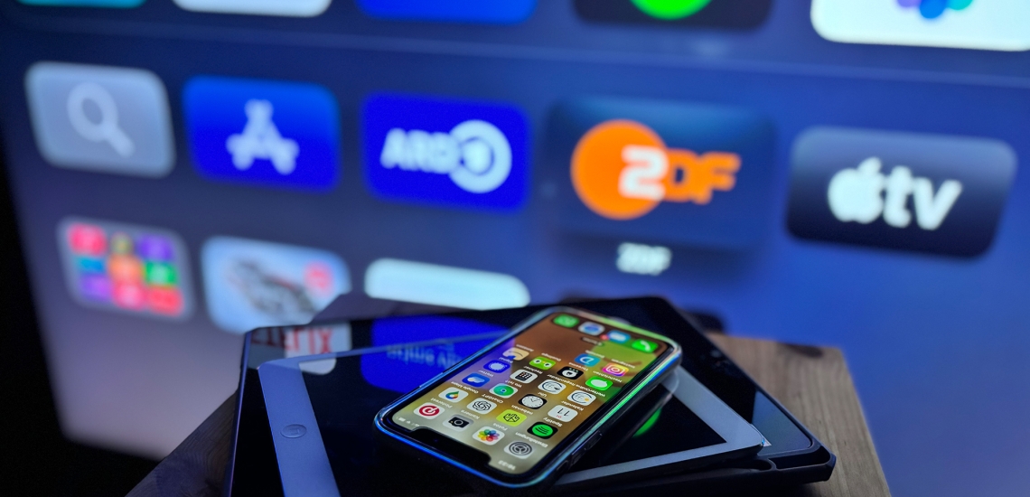 Gestapelte Tablets und ein Handy vor einem TV-Screen mit Streaming-Apps.