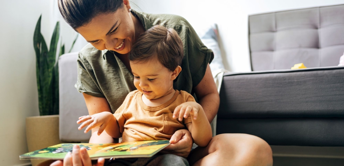 Mutter und kleines Kind sitzen fröhlich vor der Couch auf dem Boden und schauen zusammen ein Bilderbuch an