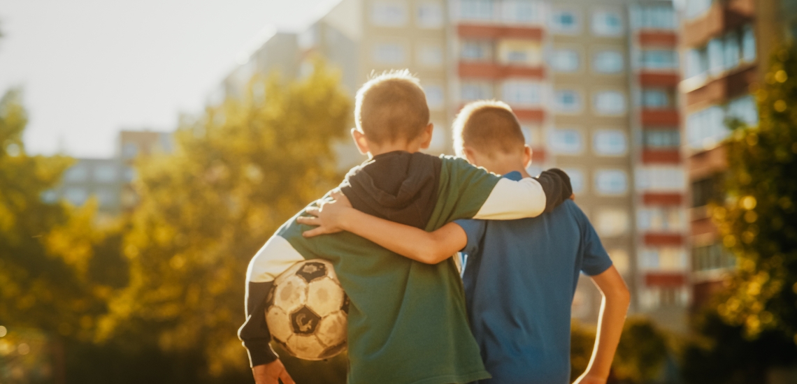 Zwei Jungen stehen vor Hochhäusern. Einer hat einen abgenutzten Fußball unter dem Arm.