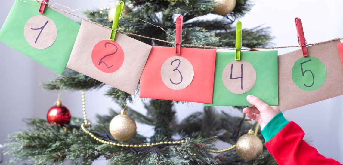 Kinderhand hält Umschläge eines hängenden Adventskalenders, im Hintergrund ein Christbaum 