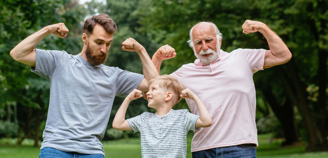 Vater, Sohn und Großvater stehen im Park und zeigen lachend ihre Muskeln
