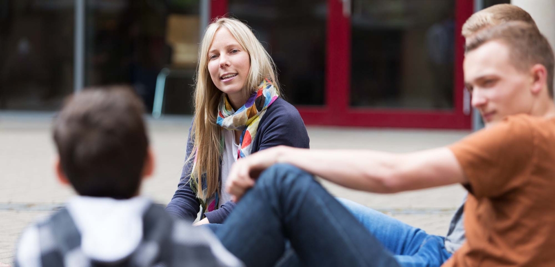 Eine junge Frau sitzt am Boden auf einem Pausenhof umringt von Kindern und Jugendlichen und hört aufmerksam zu.