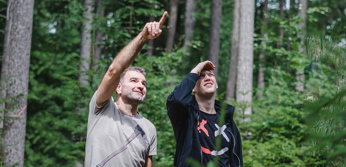 Ein Pädagoge steht neben einem Jugendlichen im Wald, beide schauen in den Himmel, der Pädagoge zeigt in die Luft.