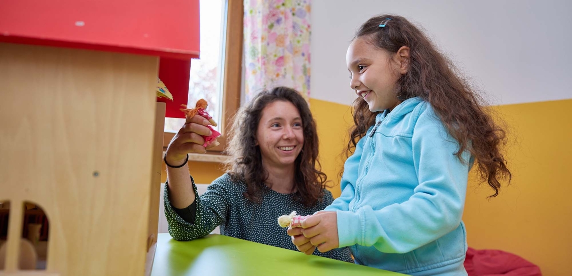 Eine Heilpädagogin und ein Mädchen spielen mit einem Puppenhaus.