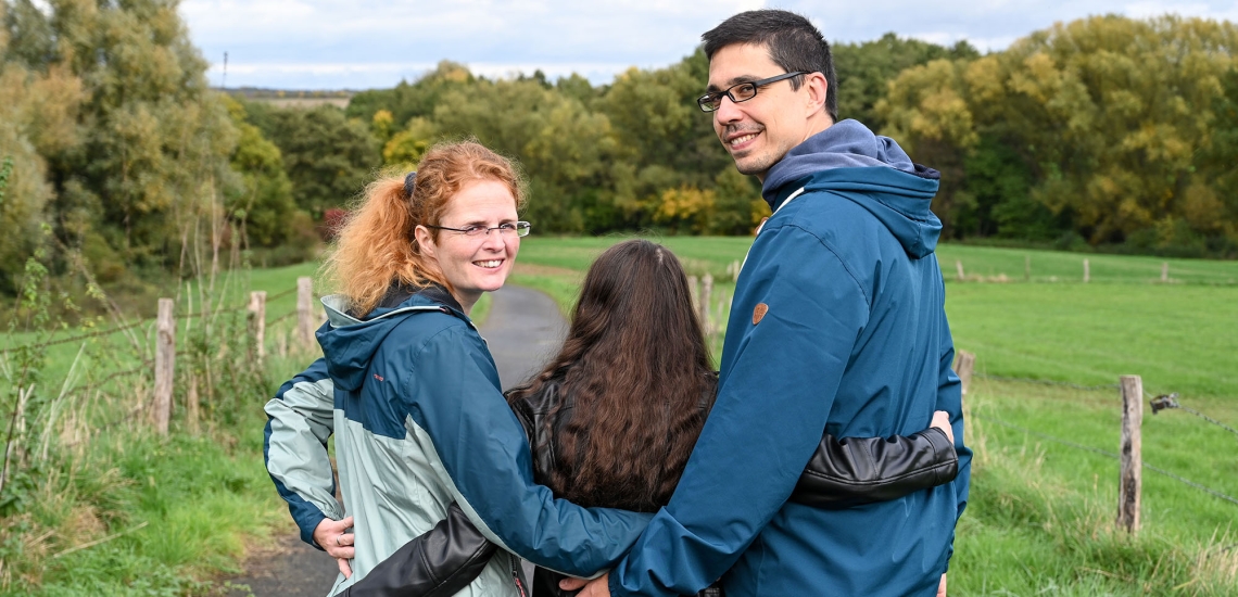 Mutter, Tochter und Kind gehen Arm in Arm spazieren. Mutter und Vater wenden ihr Gesicht zur Kamera.