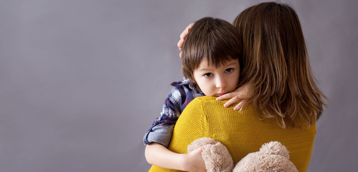 Eine Mutter hält ihren Sohn auf dem Arm. Sie ist dem Bild abgewandt, er hält seinen Teddy fest und schaut in die Kamera.