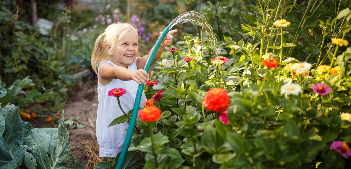 Ein kleines Mädchen steht mitten im Garten, lacht und gießt mit dem Gartenschlauch die Blumen.