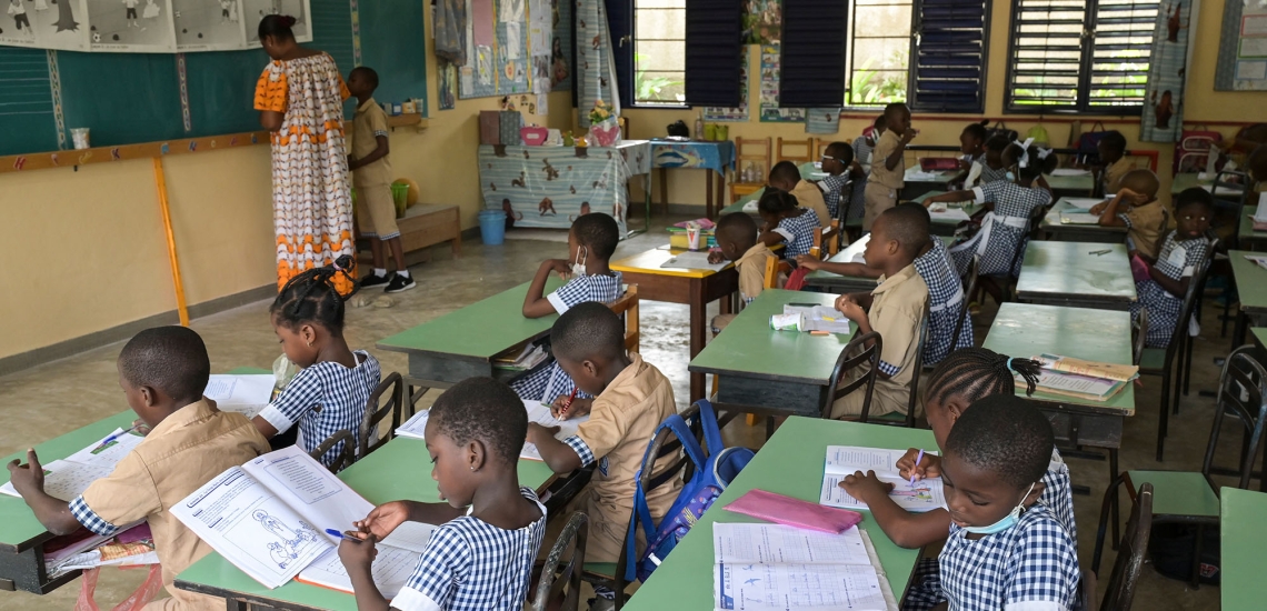 Kinder sitzen in einer Grundschulklasse, an der Tafel steht die Lehrerin.