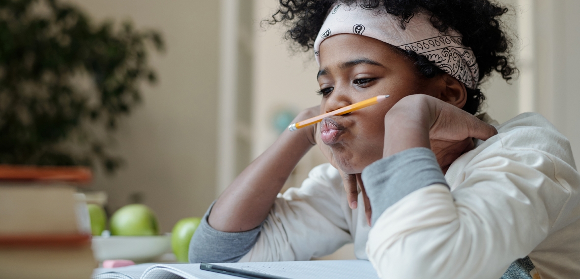 Ein Kind macht Hausaufgaben, hat seinen Bleistift zwischen Lippe und Nase eingeklemmt und schaut gelangweilt in sein Heft.
