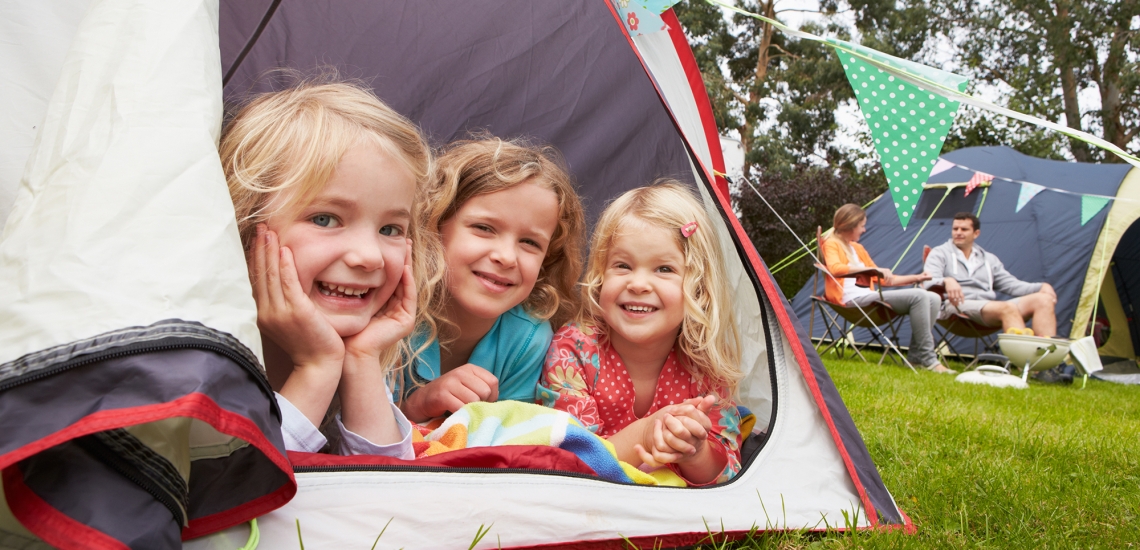 Drei kleine Mädchen liegen im Zelt und schauen lächelnd heraus. Im Hintergrund sieht man die Eltern auf Klappstühlen sitzen.