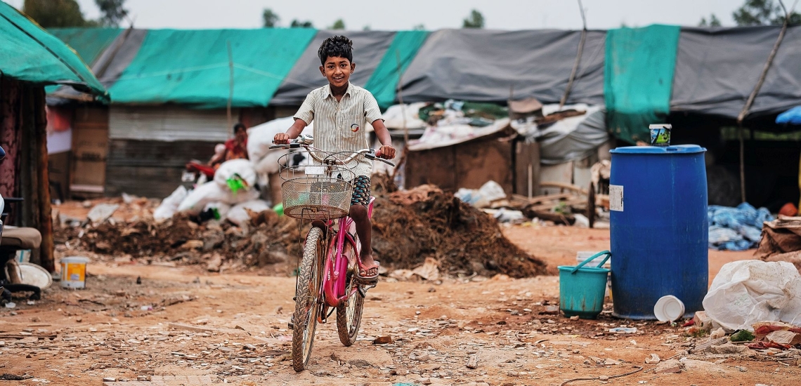 Junge fährt lachend auf sandigem Weg vor Hütten auf einem Fahrrad