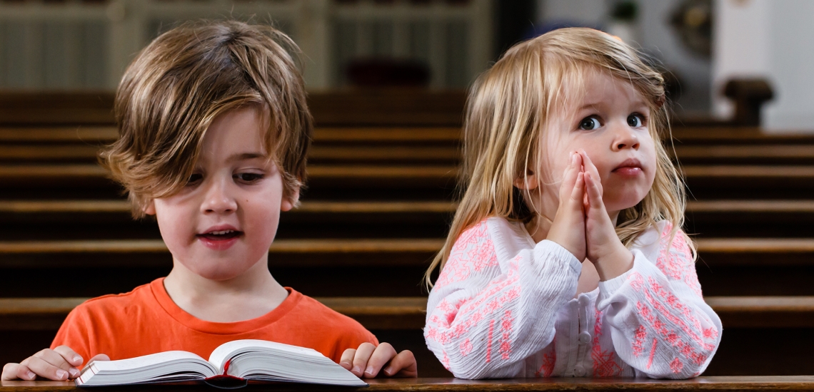 Ein Junge und ein Mädchen knien in einer Kirchenbank. Der Junge schaut ins Gotteslob, das Mädchen hat die Hände gefaltet.