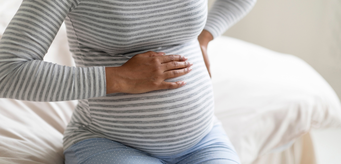 Eine Frau hält sich ihren runden Babybauch, die zweite Hand stützt den Rücken.