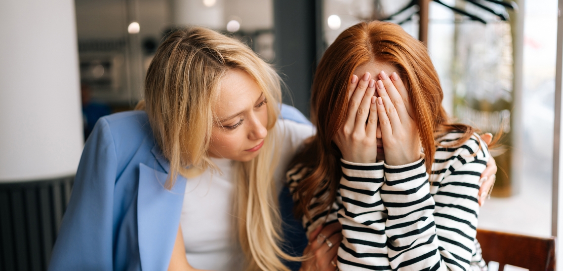 Zwei Frauen sitzen in einem Café. Eine Frau hält sich die Hände vor die Augen, die andere nimmt sie in den Arm.