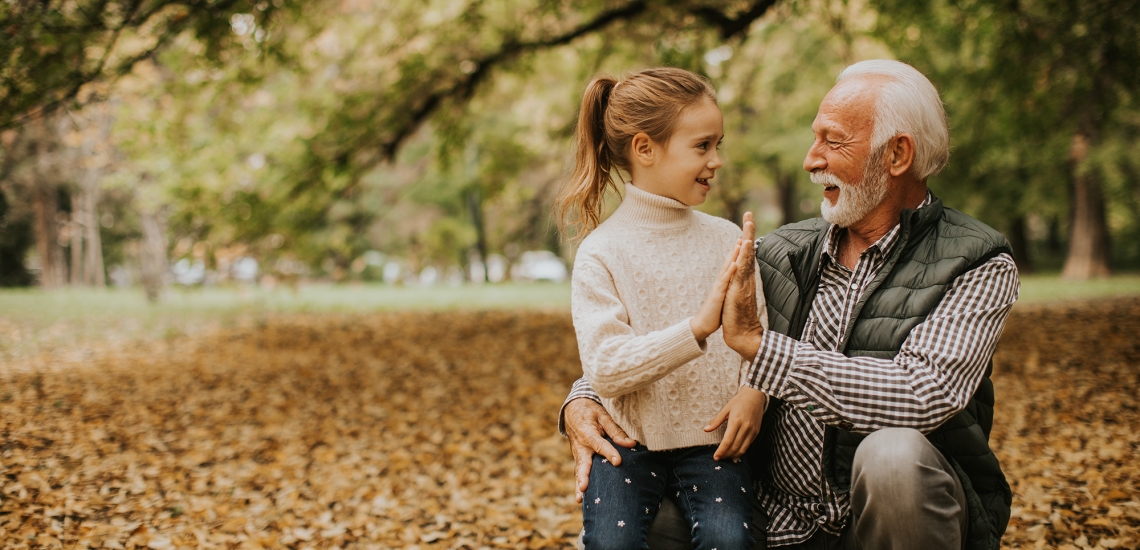 Ein Opa hält seine Hand an die seiner Enkelin. Sie befinden sich in einem herbstlichen Wald.