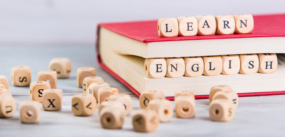 Buchstabenwürfel, die teils auf dem Tisch verstreut sind und die auf und in einem Buch so liegen, dass sie "Learn English" ergeben.