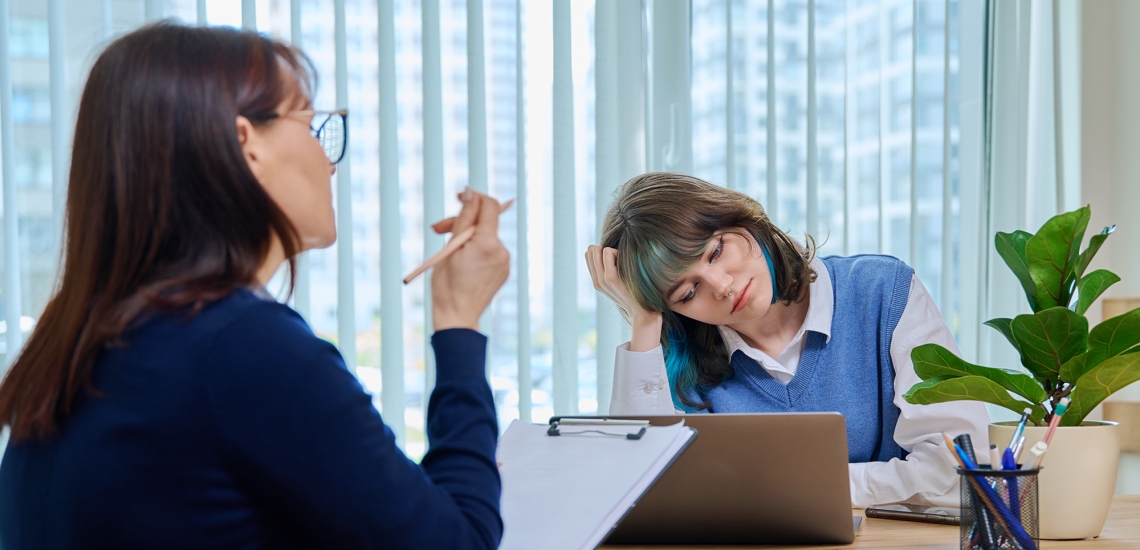 Eine Frau und ein junges Mädchen sitzen bei einem Gespräch am Tisch. Das Mädchen macht ein trauriges Gesicht und stützt ihren Kopf ab.