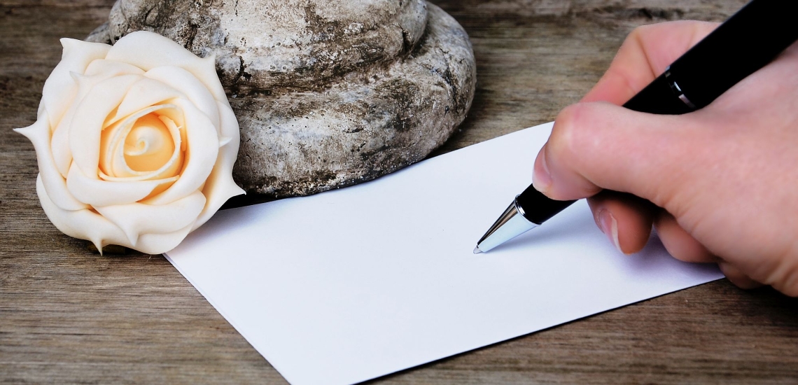 Hand mit Stift schreibt Trauerkarte, daneben Stein und cremeweiße Rose 