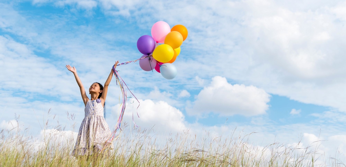 Kind in Sommerkleid auf Wiese hält Luftballons hoch in die Luft und streckt die Hände zum Himmel 