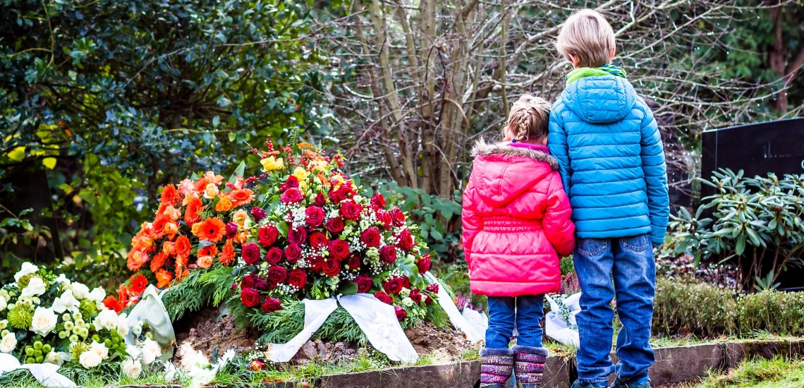 Junge und Mädchen stehen an frisch aufgeschüttetem Grab mit Kränzen und halten sich an den Händen 