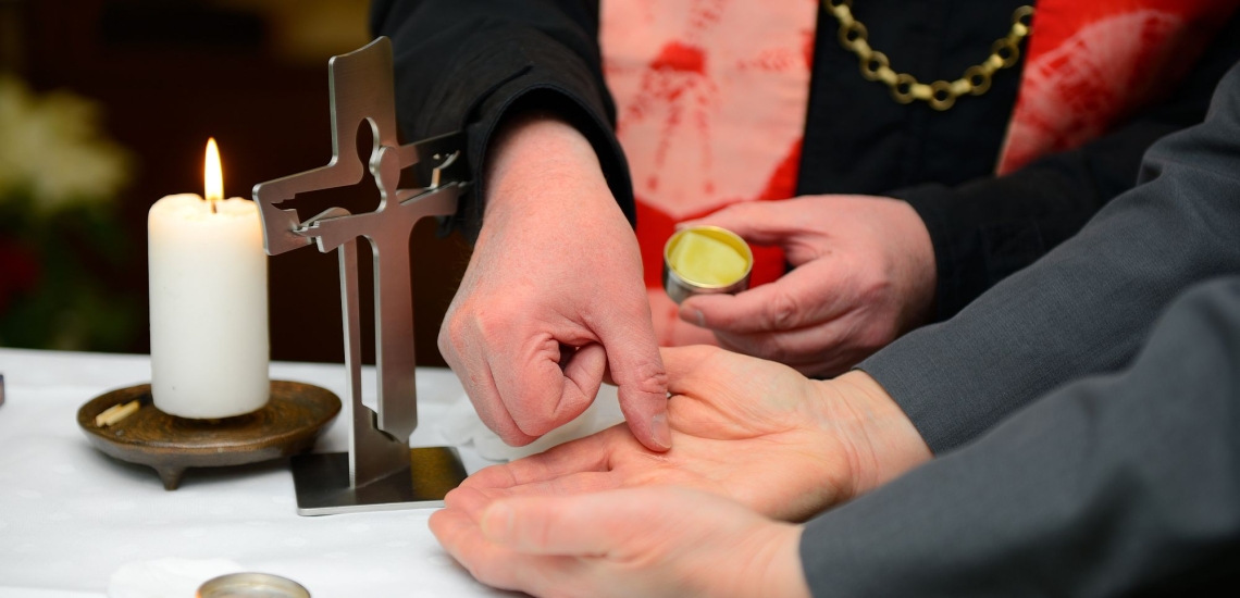 Ein Priester salbt einen Kranken mit Krankenöl, daneben ein Kreuz und eine brennende Kerze 
