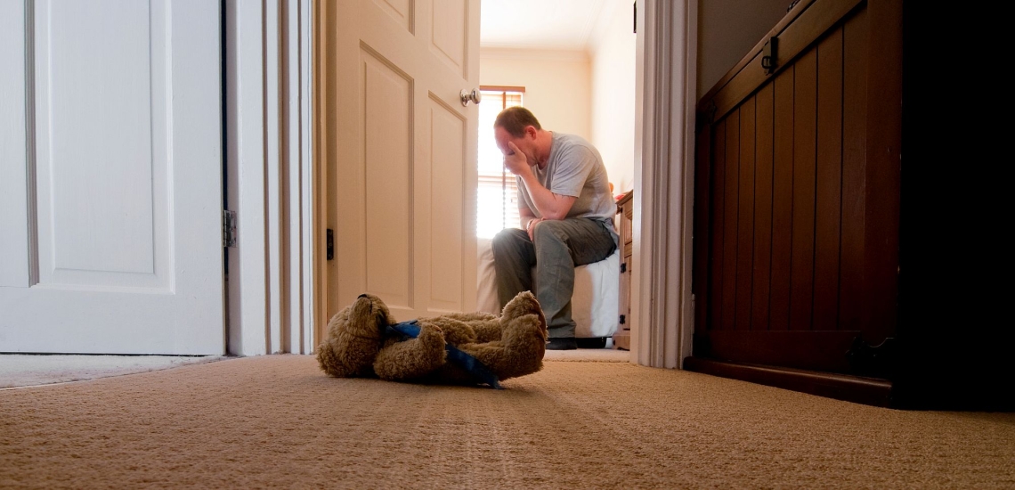 Trauernder Vater sitzt im Kinderzimmer, vor dem Zimmer liegt ein Teddybär auf dem Teppich 