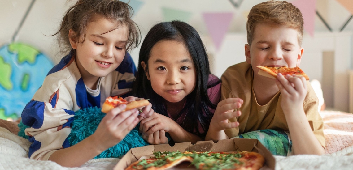 Drei Kinder liegen zusammen auf Bett und essen Pizza aus Karton 