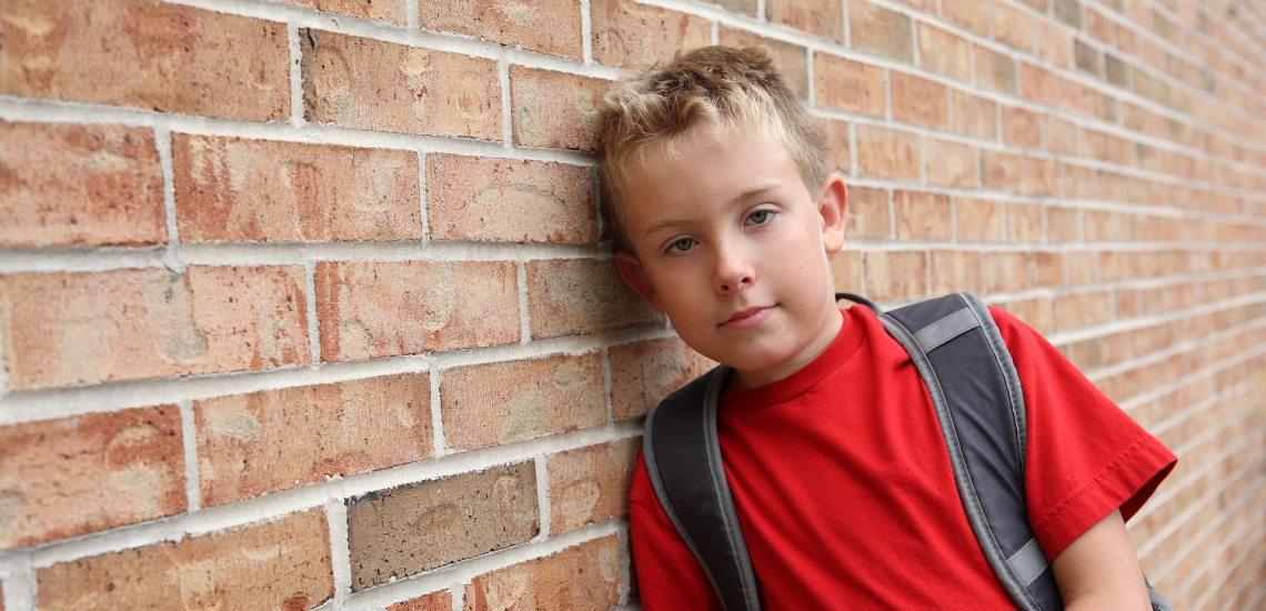 Junge mit Schulrucksack lehnt schüchtern an einer Mauer 