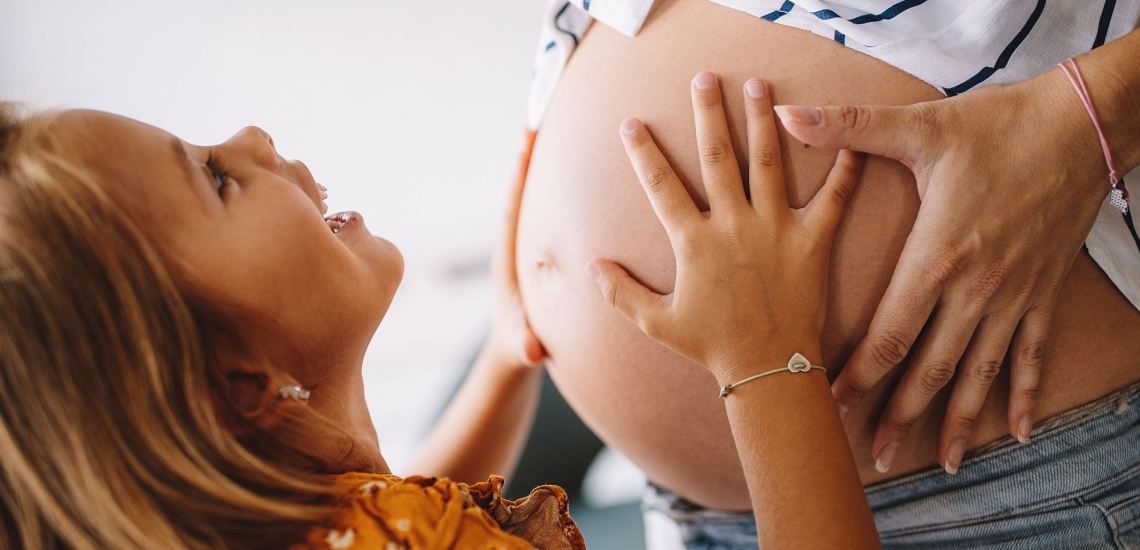 Kind legt Hände auf Bauch einer schwangeren Frau und schaut fragend und glücklich nach oben 