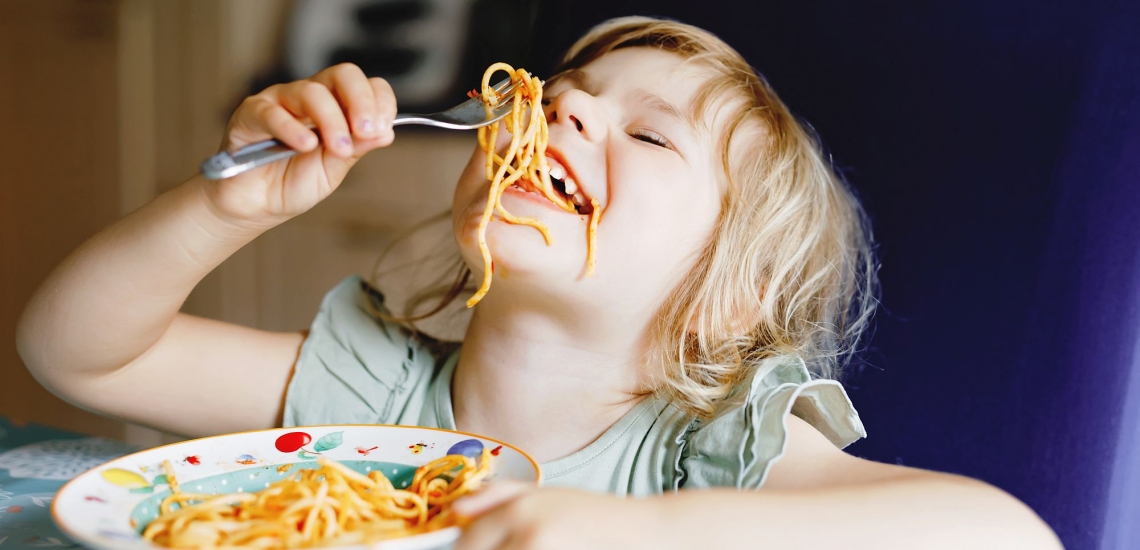 Kind lässt Spaghetti mit Soße von der Gabel in den Mund hängen 