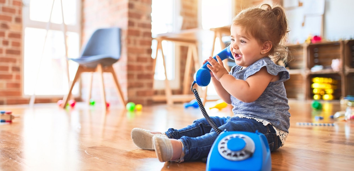 Kleinkind sitzt auf Boden und spricht begeistert in ein Spielzeugtelefon 