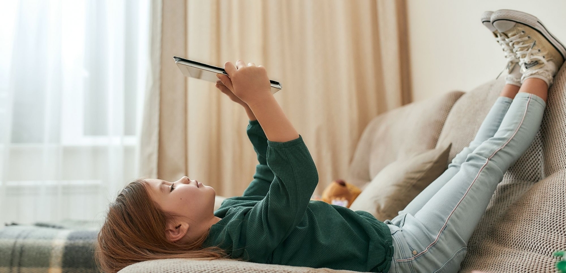 Kind liegt mit den Füßen nach oben auf dem Sofa und schaut auf ein Tablet